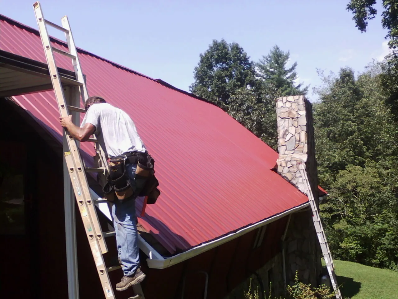 Roofer on a ladder beside a red metal roof and stone chimney — Soffit & Fascia Repair by Roofing Brothers in Mascotte
